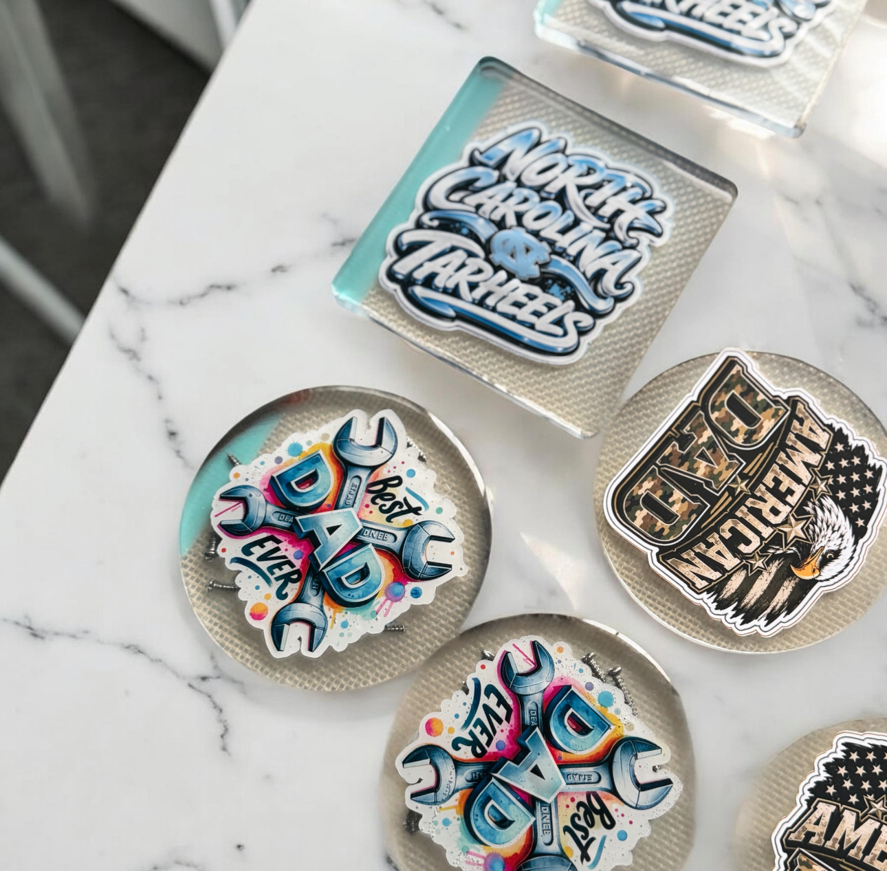 Flat lay of round resin coasters on a marble surface featuring Carolina Panthers and Best Dad Ever designs with wrench graphics in blue, pink and yellow.